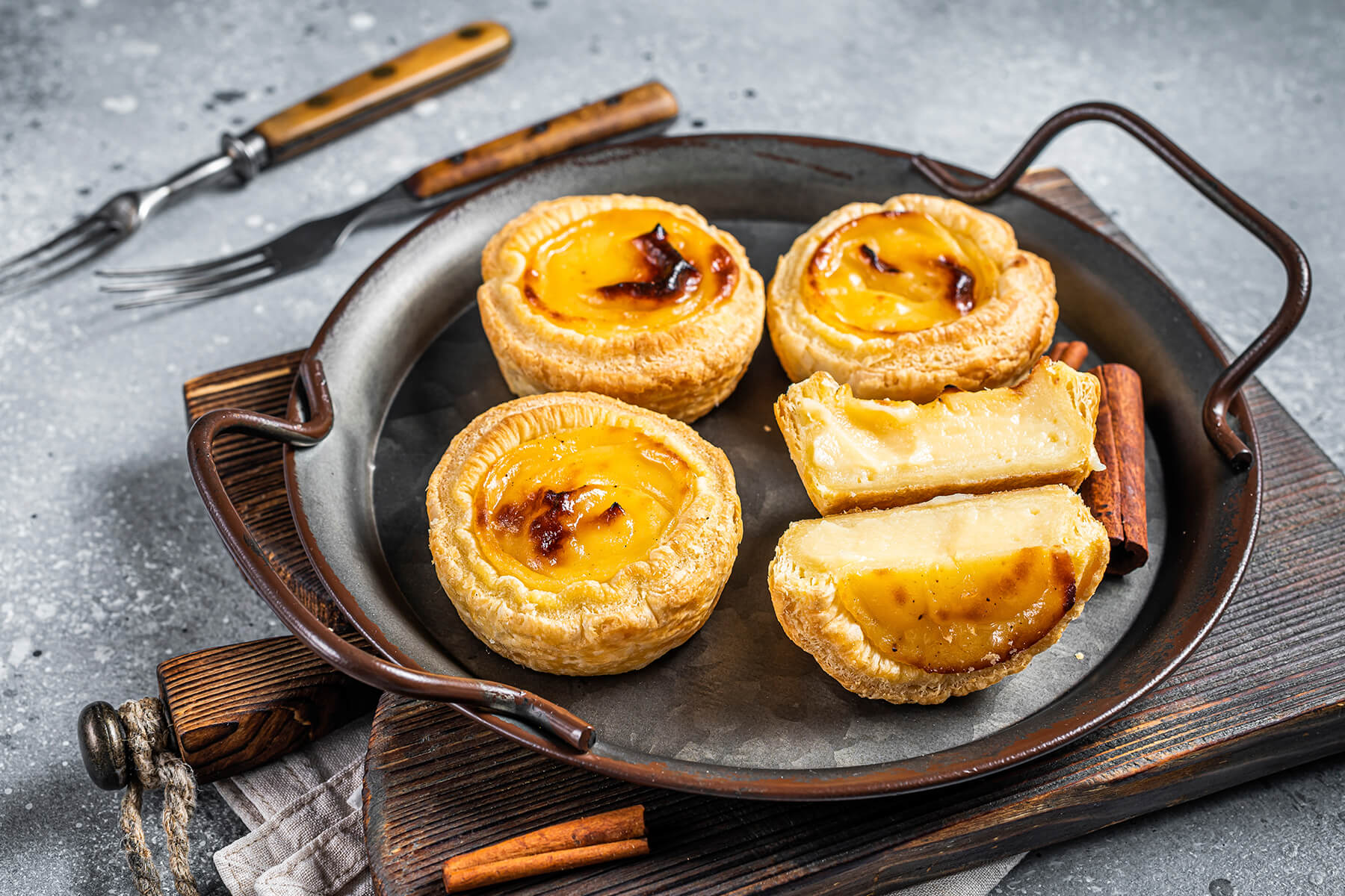 Aufgeschnittenes Pastel de Nata auf einem dunklen Teller, daneben ganze Törtchen und Zimtstangen