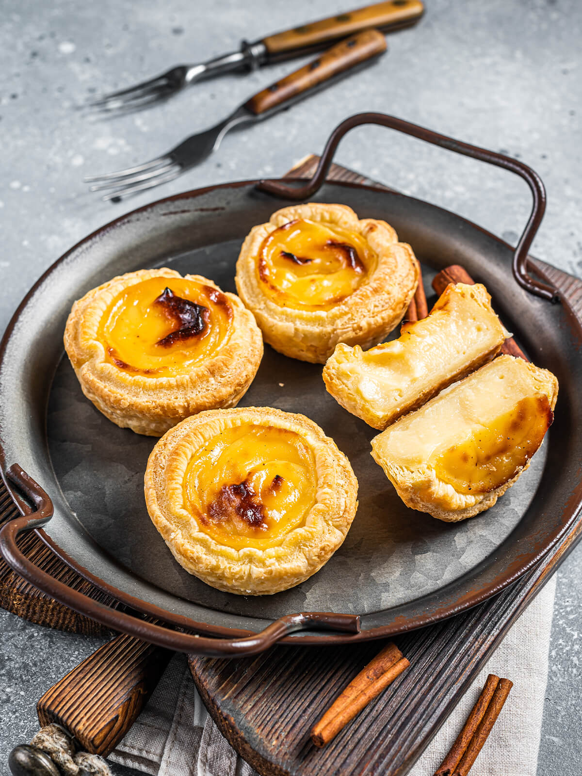 Drei ganze und ein aufgeschnittenes Pastel de Nata in einer dunklen Servierpfanne mit Zimtstangen