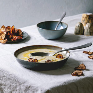 Cremige Pastinaken-Suppe in dunkler Schale mit Olivenöl-Drizzle, Kräutern und frischen Pastinaken auf grauem Untergrund