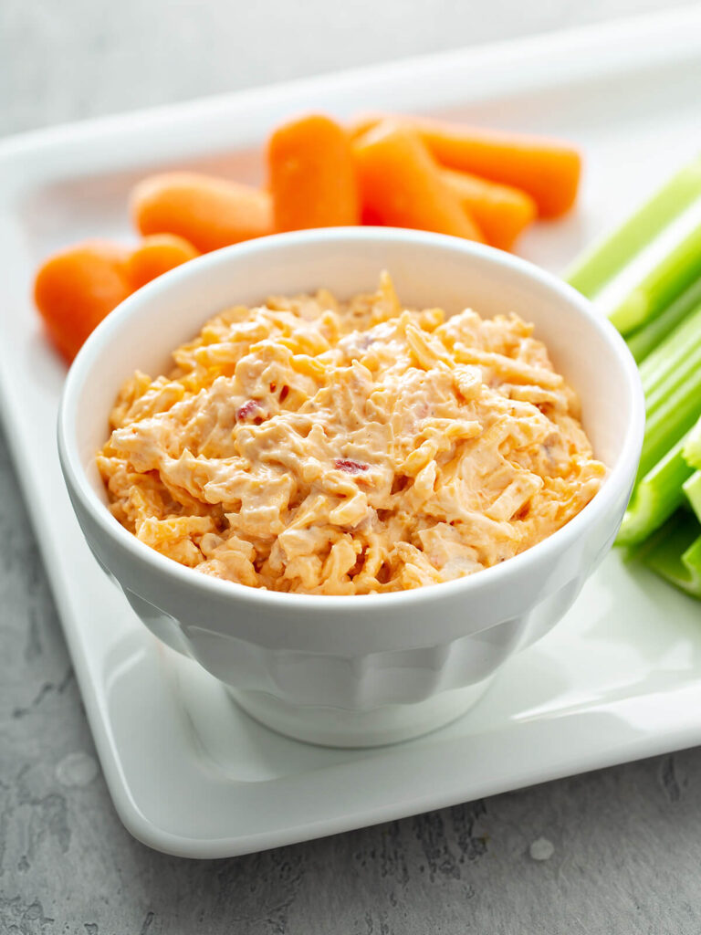 Cremiger Pimento Cheese Dip in weißer Schale mit geriebenem Cheddar, sichtbaren Pimento-Stückchen, Selleriestangen und Babykarotten