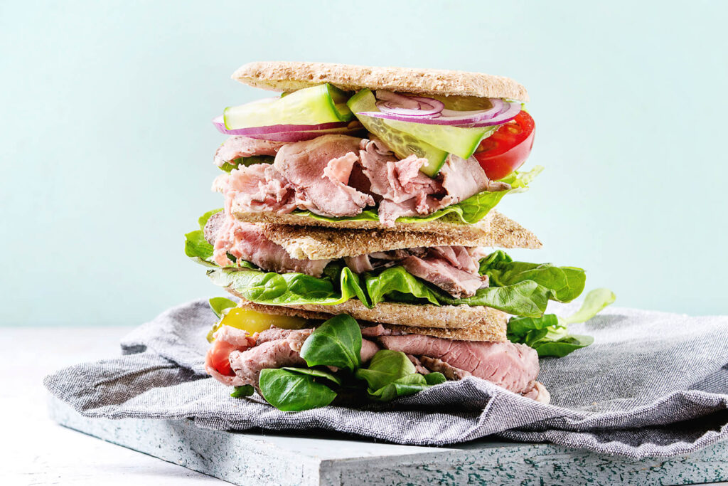Hoch gestapeltes Roastbeef-Sandwich mit rosa Roastbeef, Salat, Gurke, Zwiebel und Tomate
