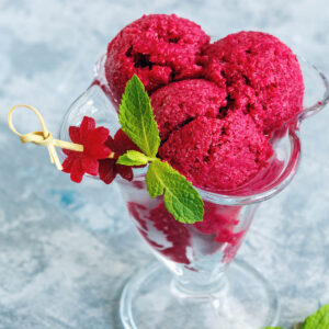 Drei dunkelrote Kugeln Rote-Bete-Sorbet in einem hohen Kristall-Glasbecher, mit frischer Minze und essbarer Blüte als Dekoration