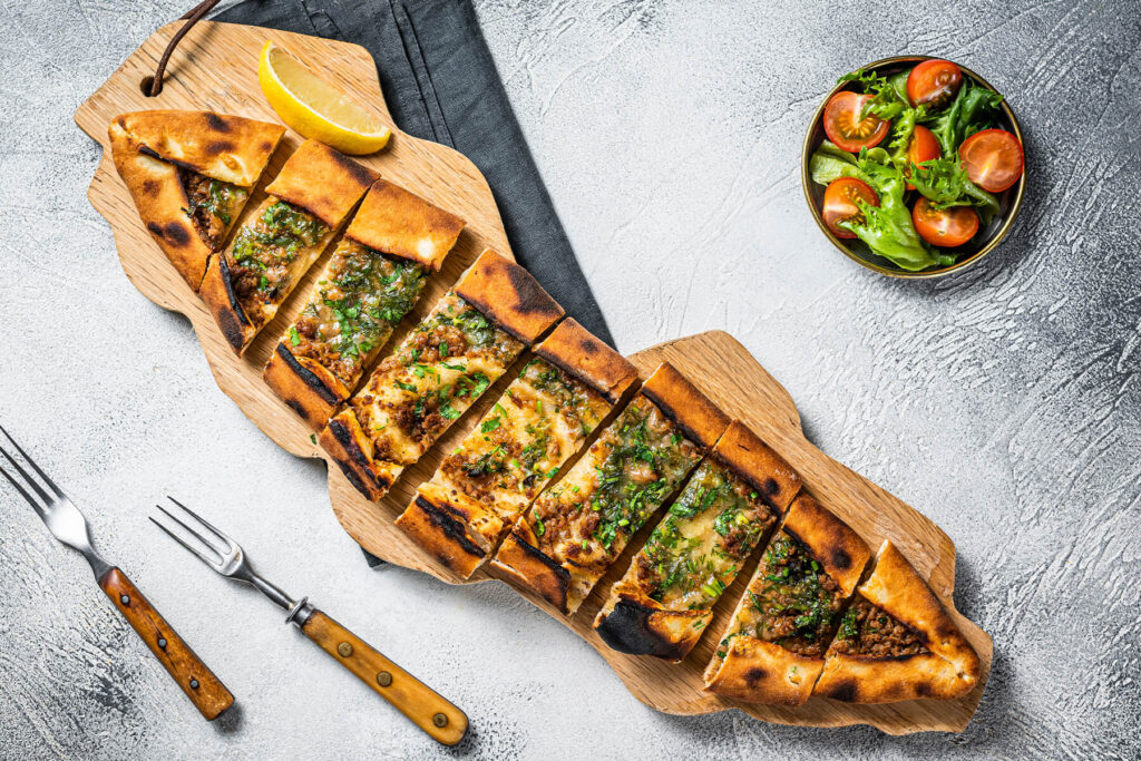 Aufgeschnittene türkische Pide mit Hackfleisch und Käse auf einem Holzbrett, daneben eine Schale grüner Blattsalat mit Kirschtomaten und eine Zitronenspalte