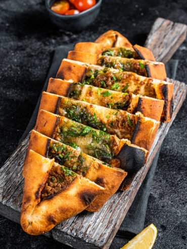 Türkische Pide mit Hackfleisch in Scheiben auf einem Holzbrett, daneben eine Schale Kirschtomaten und eine Zitronenspalte
