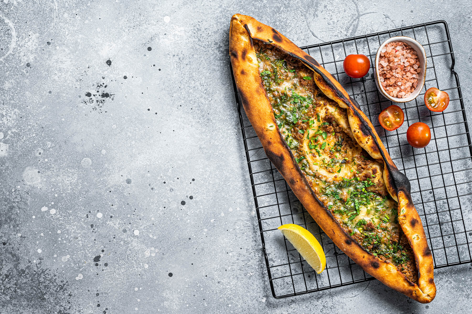 Frisch gebackene türkische Pide mit Hackfleisch, geschmolzenem Käse und frischen Kräutern auf einem Gitter, daneben Kirschtomaten und eine Schale mit rosa Salz