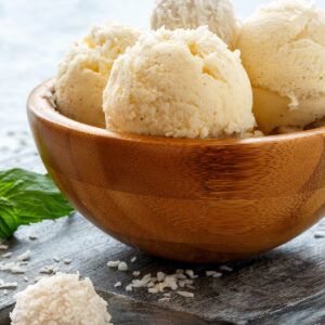 Cremige Kugeln veganes Kokos-Vanille-Eis in einer Holzschale mit Kokosraspeln, weißen Kokoskugeln und frischer Minze