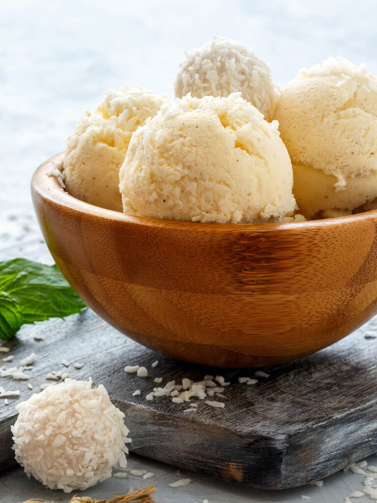 Cremige Kugeln veganes Kokos-Vanille-Eis in einer Holzschale mit Kokosraspeln, weißen Kokoskugeln und frischer Minze