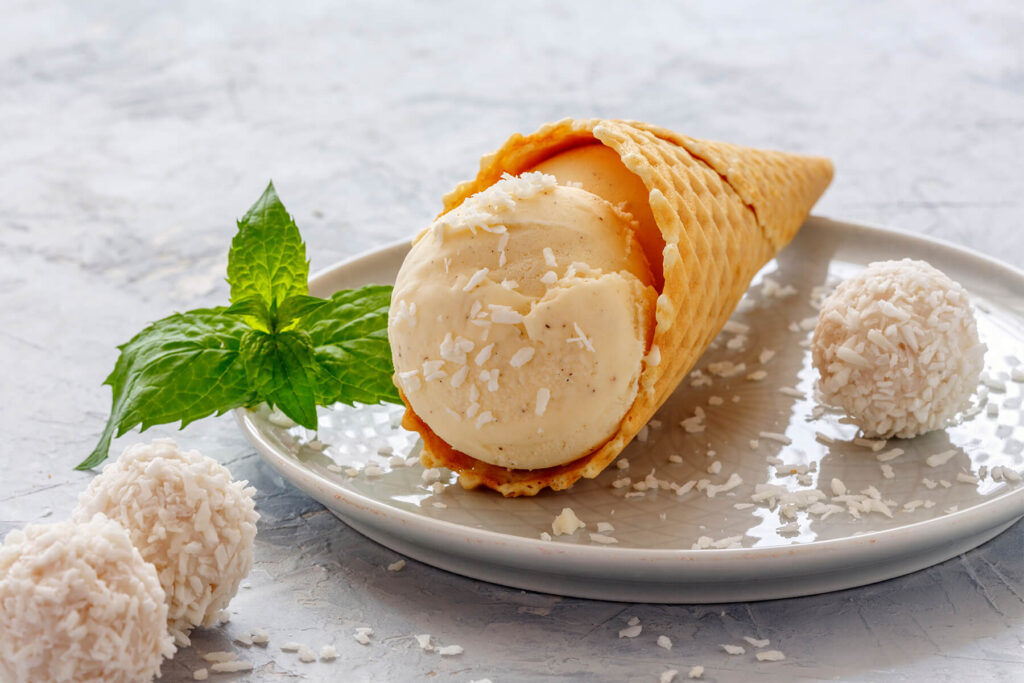 Cremige Kugeln veganes Kokos-Vanille-Eis in einer Holzschale mit Kokosraspeln, Kokostrüffeln und Minze