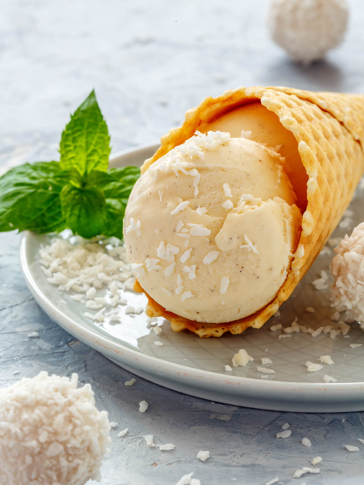 Eine Kugel veganes Kokos-Vanille-Eis in einer Waffeltüte auf einem Teller, garniert mit Kokosraspeln, weißen Kokoskugeln und frischer Minze