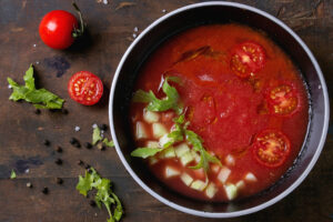Tiefrote Wassermelonen-Tomaten-Gazpacho in dunkler Schale mit Gurkenwürfeln, Rucola und halbierten Kirschtomaten