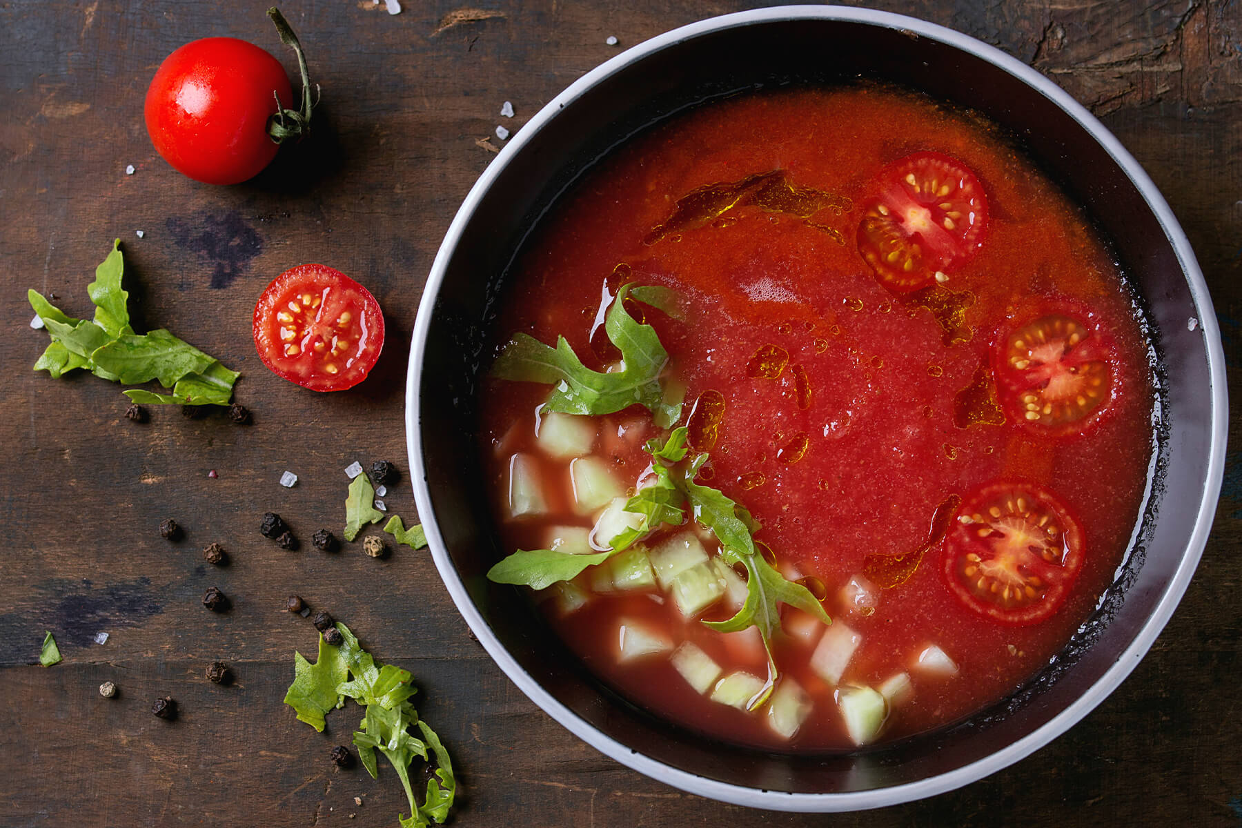 Tiefrote Wassermelonen-Tomaten-Gazpacho in dunkler Schale mit Gurkenwürfeln, Rucola und halbierten Kirschtomaten