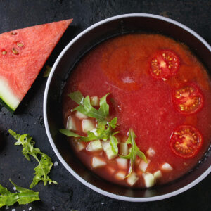 Kalte Wassermelonen-Tomaten-Gazpacho in dunkler Schale mit Gurkenwürfeln, Rucola und halbierten Kirschtomaten