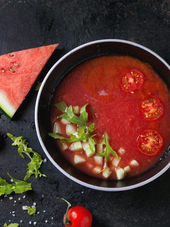 Kalte Wassermelonen-Tomaten-Gazpacho in dunkler Schale mit Gurkenwürfeln, Rucola und halbierten Kirschtomaten