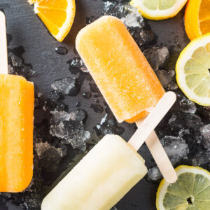 Orangefarbene und hellgelbe Zitrus-Stieleis-Portionen auf einem Schieferbrett mit Crushed Ice und Zitrusscheiben