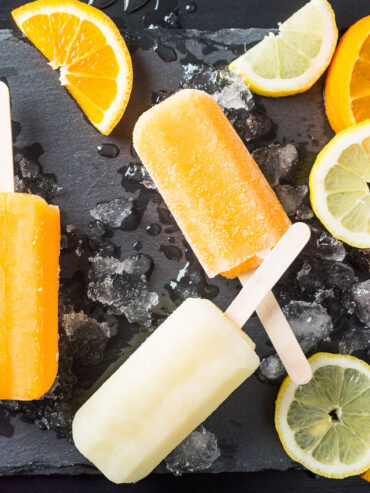 Orangefarbene und hellgelbe Zitrus-Stieleis-Portionen auf einem Schieferbrett mit Crushed Ice und Zitrusscheiben