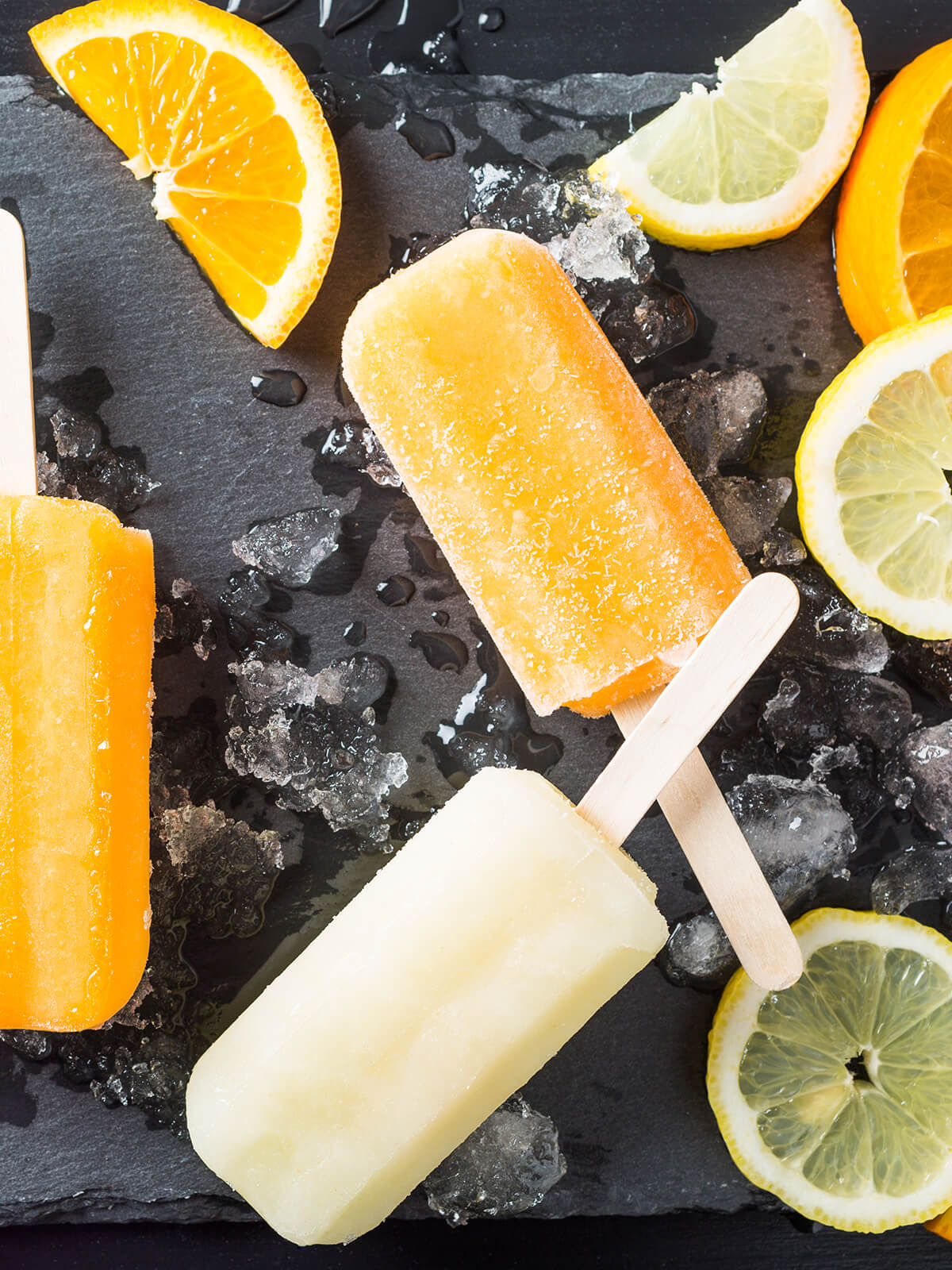 Orangefarbene und hellgelbe Zitrus-Stieleis-Portionen auf einem Schieferbrett mit Crushed Ice und Zitrusscheiben