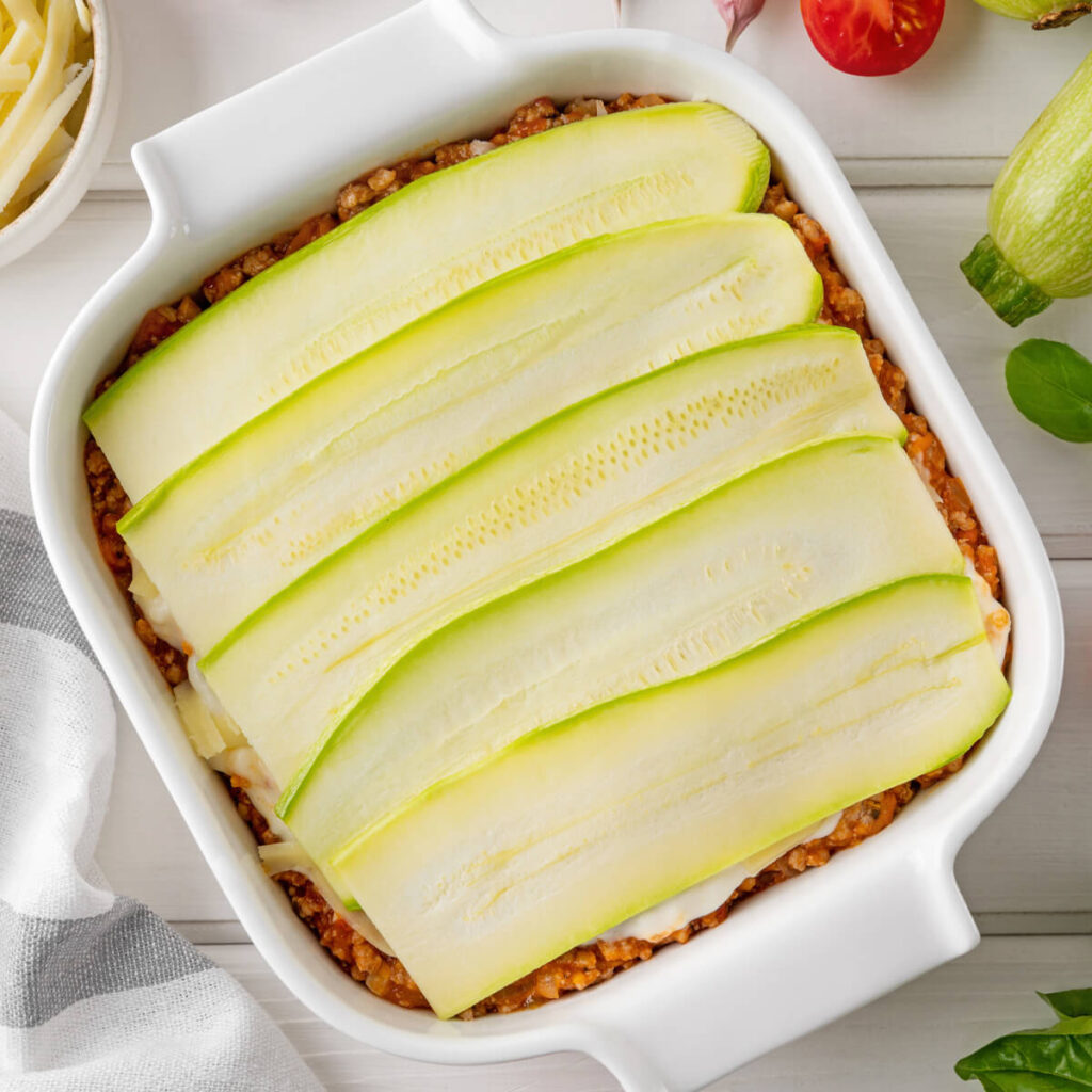 Zucchini-Lasagne beim Schichten – letzte Lage aus rohen Zucchinistreifen auf der Hackfleischsauce in der Auflaufform
