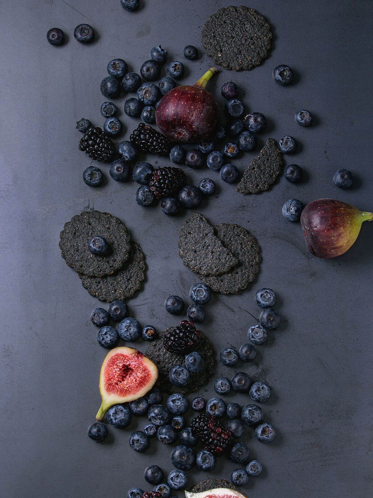 Runde schwarze Saaten-Cracker mit sichtbaren Sesamkörnern, Heidelbeeren, Brombeeren und Feigen auf dunklem Untergrund
