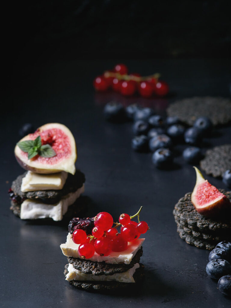 Schwarze Saaten-Cracker mit Aktivkohle, Weichkäse-Scheiben, Feigen und Beeren vor dunklem Hintergrund