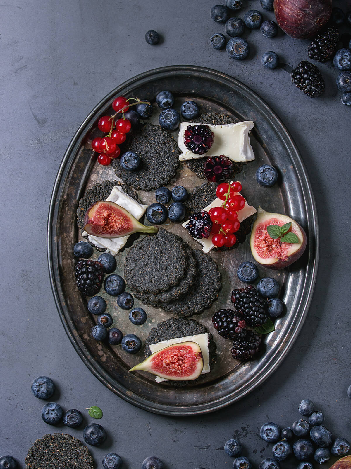 Schwarze Saaten-Cracker mit Weichkäse-Scheiben, halbierten Brombeeren, Johannisbeeren und Feigen auf einem matten Metalltablett