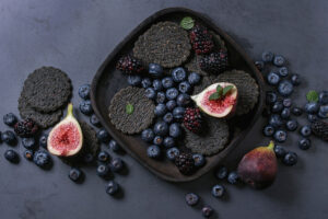 Schwarze Saaten-Cracker in dunkler Holzschale mit Brombeeren, Heidelbeeren und halbierten Feigen
