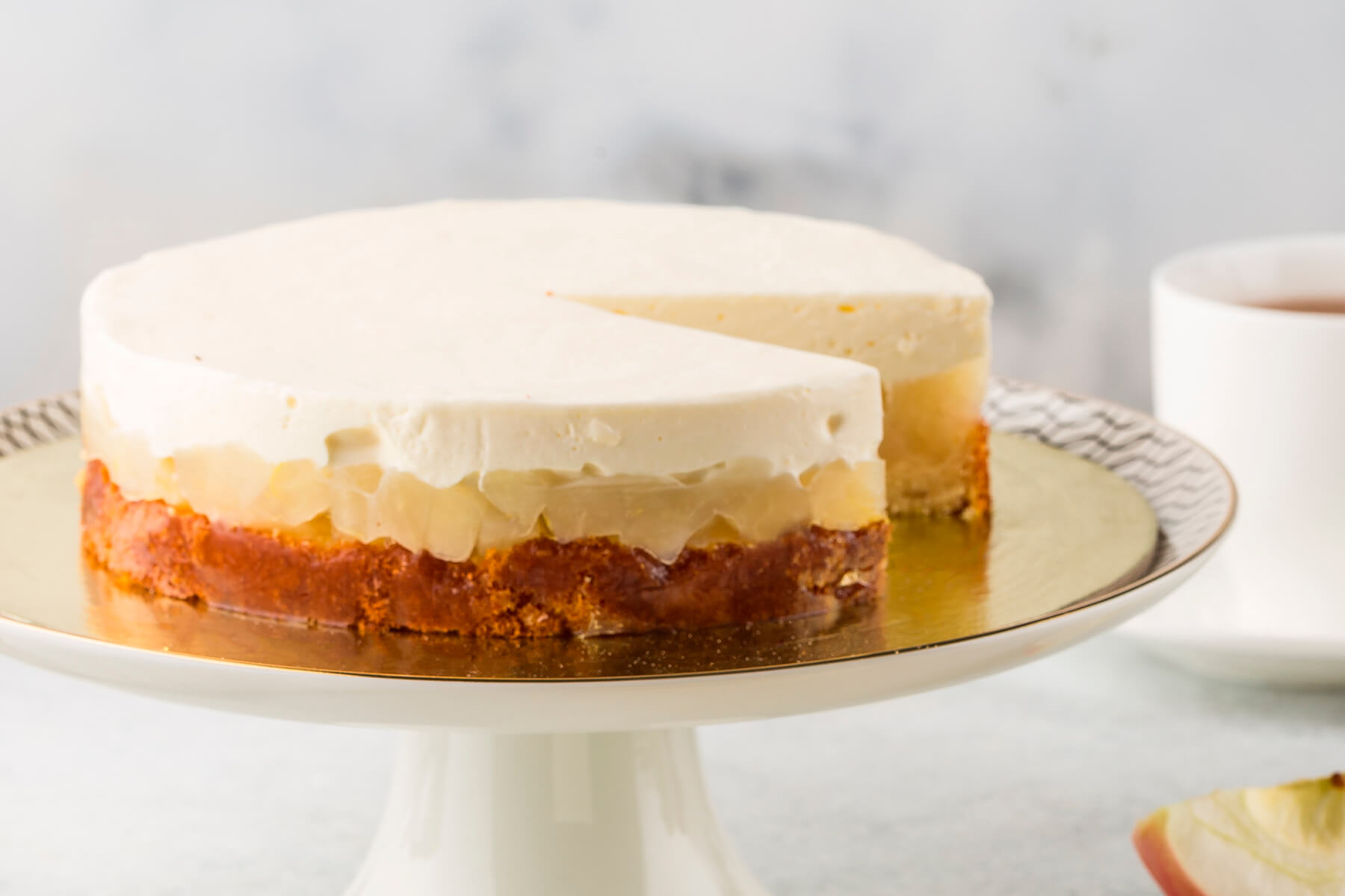 Ganzer Apfel-Cheesecake mit angeschnittenem Stück auf goldenem Tortenboden, daneben eine Teetasse