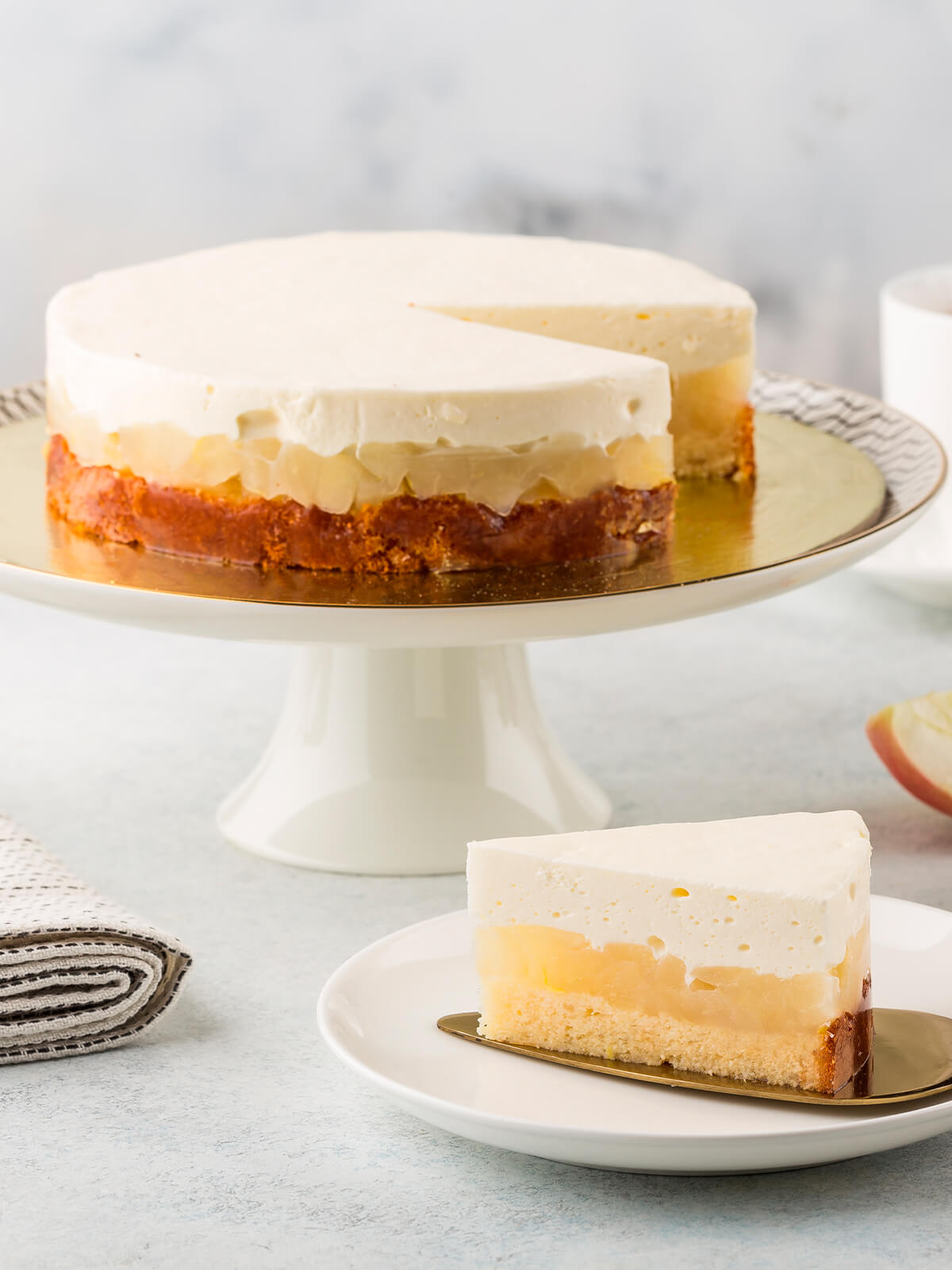 Apfel-Cheesecake aus Biskuit, Apfel-Gelee und luftiger Frischkäse-Creme auf weißem Tortenständer mit Teetasse