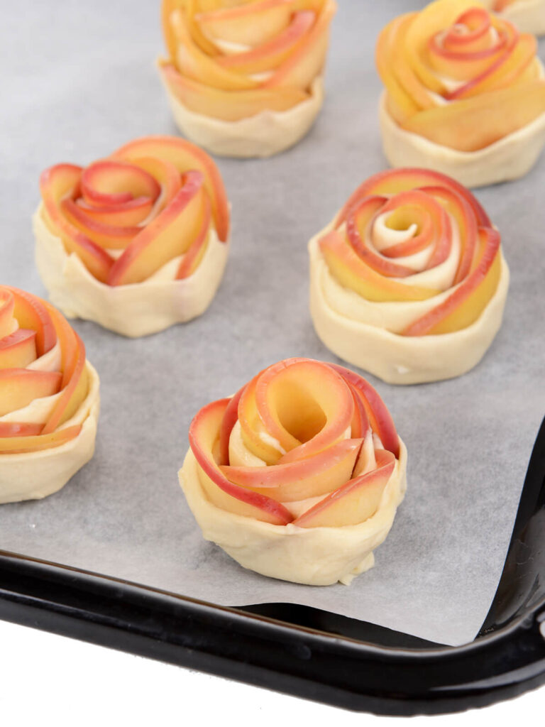 Mehrere aufgerollte rohe Apfel-Rosen auf einem Backblech vor dem Backen