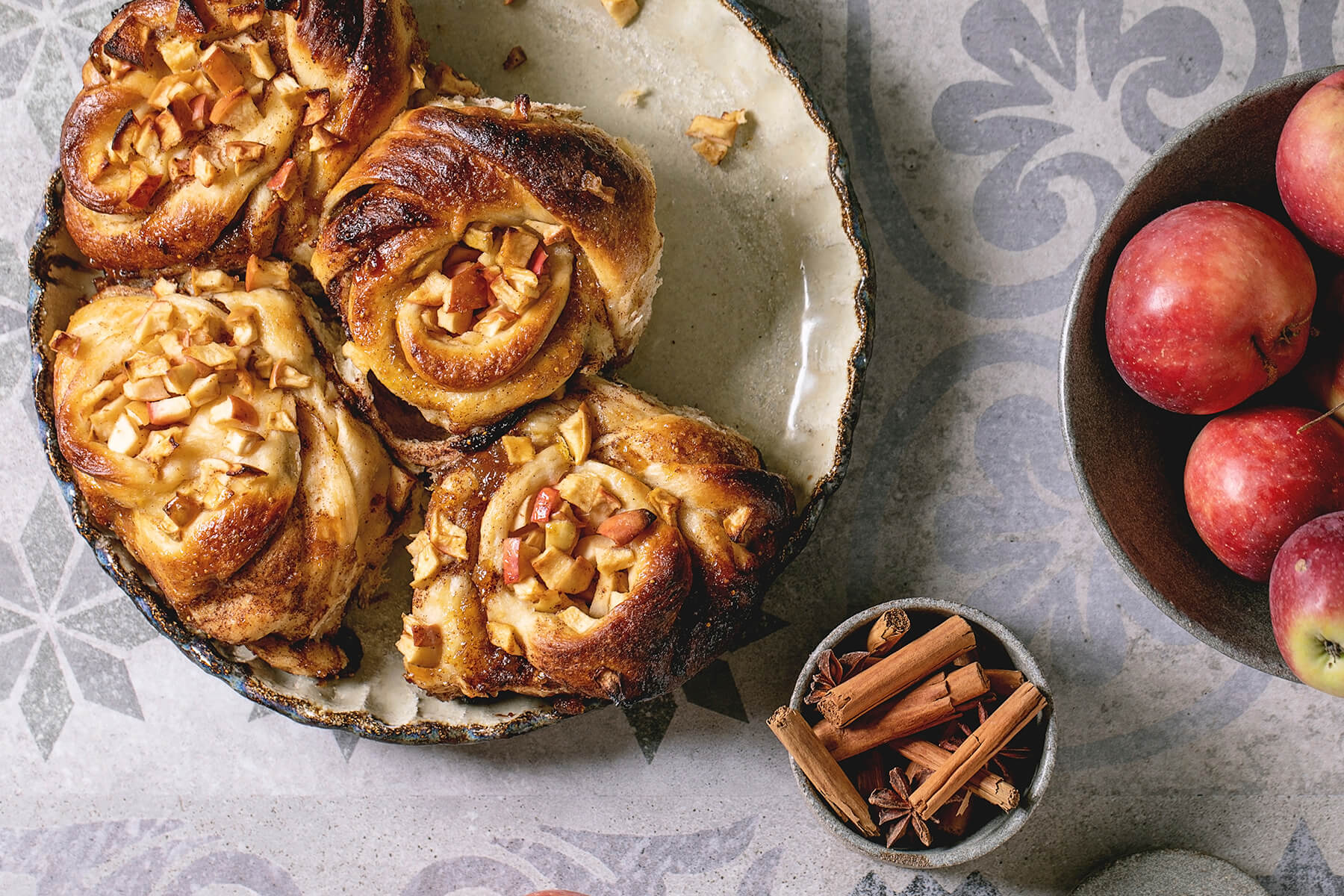 Angebrochenes Apfel-Zimt-Pull-Apart-Brot in dunkler Keramikform mit einer Schale roter Äpfel und Zimtstangen daneben