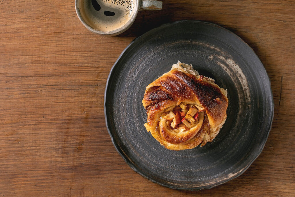 Einzelne Apfel-Zimt-Spirale mit Apfelwürfeln auf einem dunklen Teller neben einer Tasse Kaffee