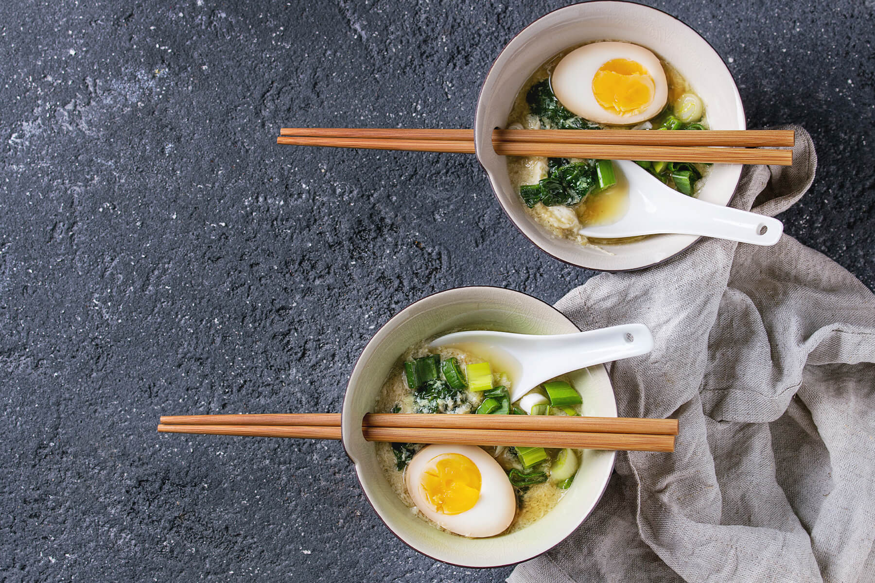Zwei Schalen asiatische Eier-Spinat-Suppe mit halbiertem Ei, Frühlingszwiebeln und Bambusstäbchen von oben