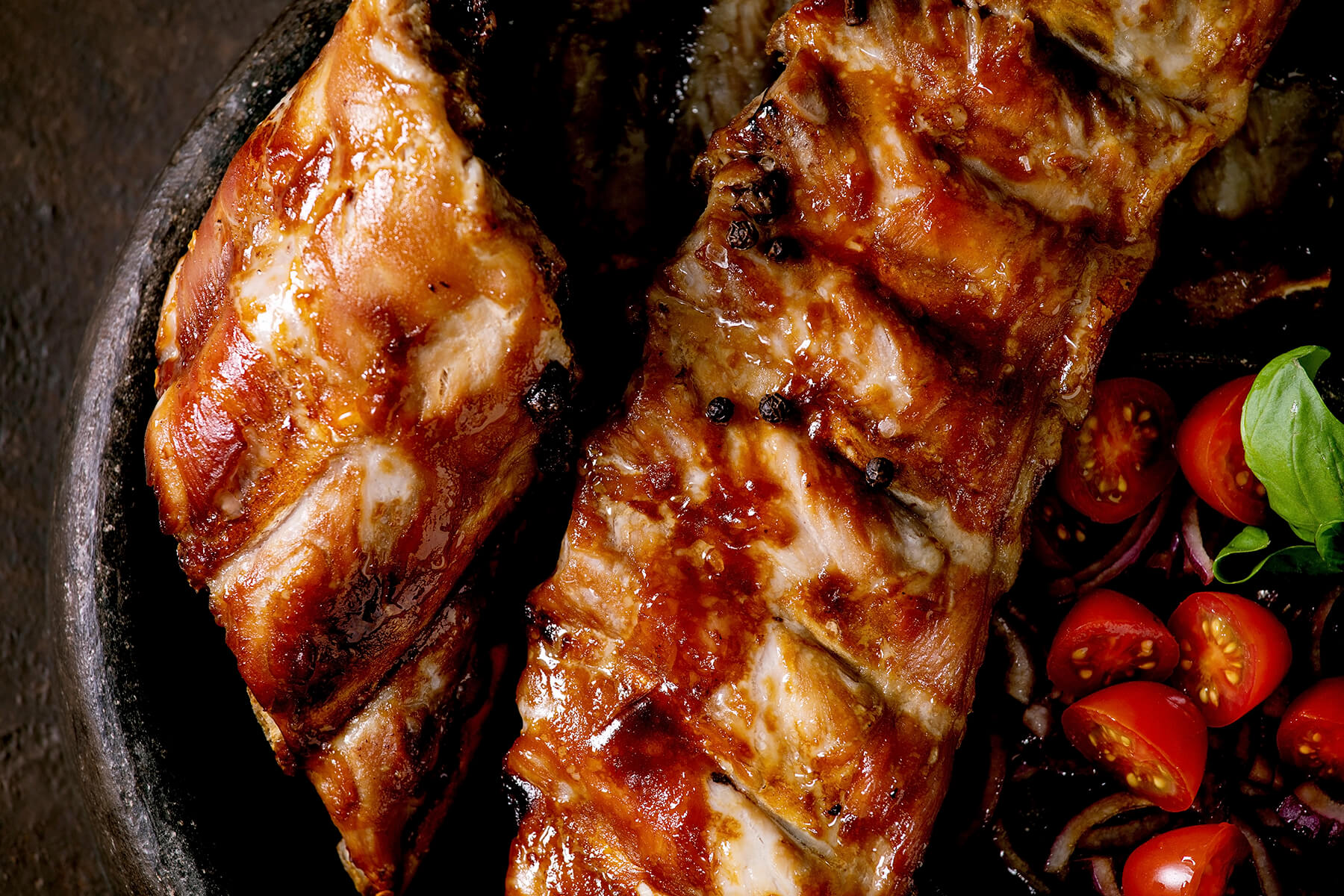 Spareribs mit dunkel glänzender, karamellisierter BBQ-Glasur, daneben halbierte Cherrytomaten mit roten Zwiebeln