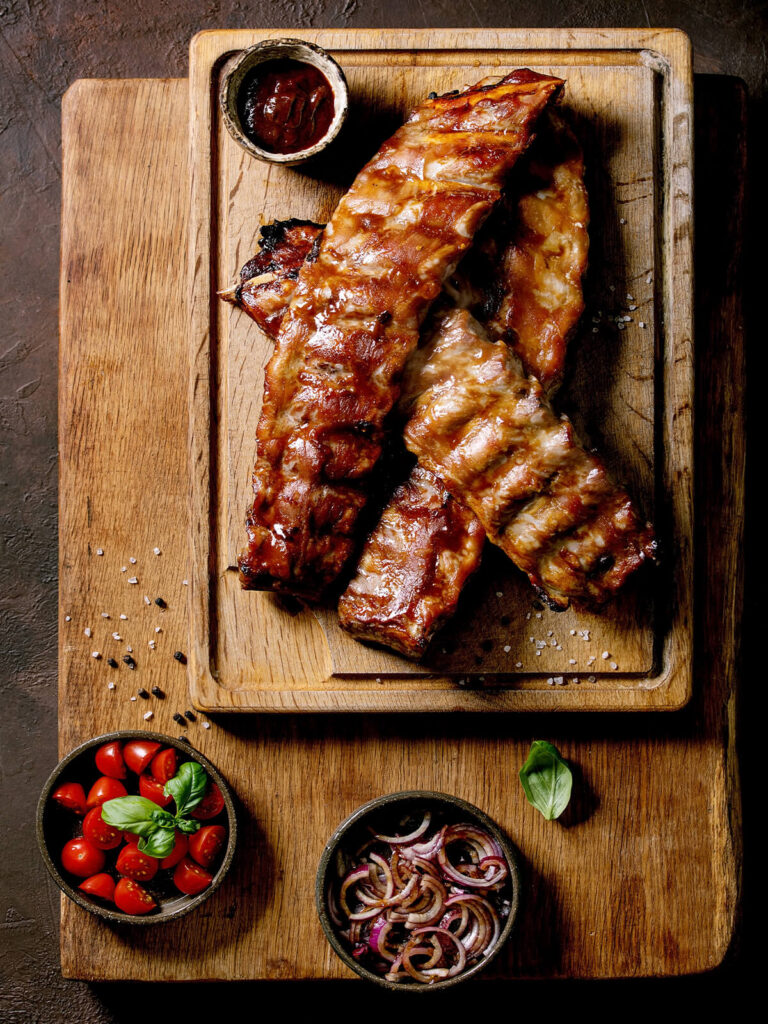 Spareribs auf einem Holzbrett mit Schälchen für BBQ-Sauce, Tomaten und rote Zwiebeln