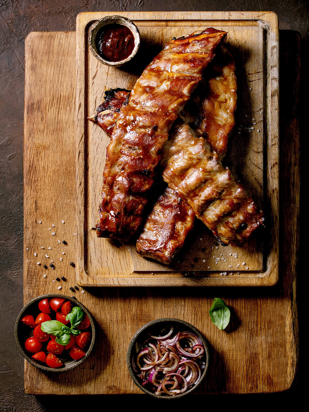Spareribs auf einem Holzbrett mit Schälchen für BBQ-Sauce, Tomaten und rote Zwiebeln