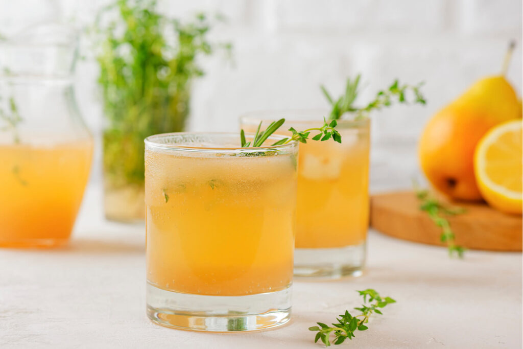 Zwei Tumbler-Gläser Birnen-Limonade mit Rosmarin und Thymian, dahinter ein Kräuterstrauß und frische Birnen