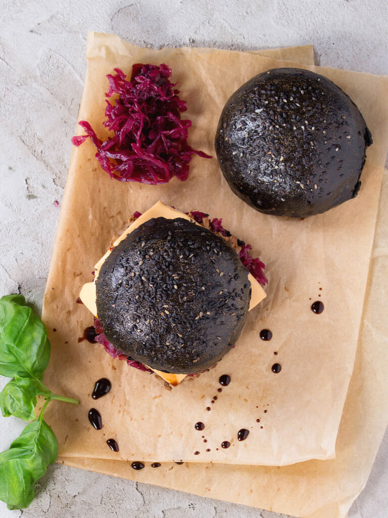 Zwei Black Burger mit Aktivkohle-Brötchen von oben, daneben frisch mariniertes pinkes Rotkraut und frisches Basilikum auf Backpapier