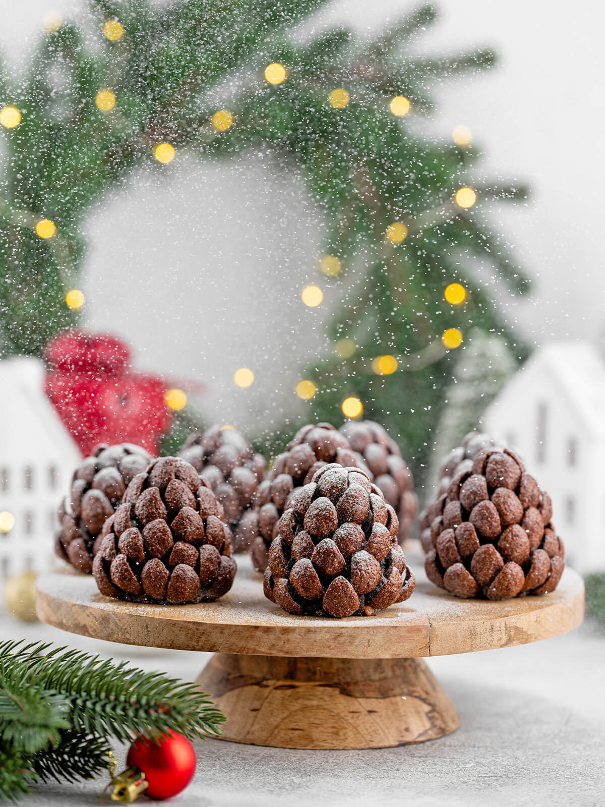 Mehrere Schoko-Tannenzapfen auf einem Holzteller vor weihnachtlicher Kulisse mit Lichterkette