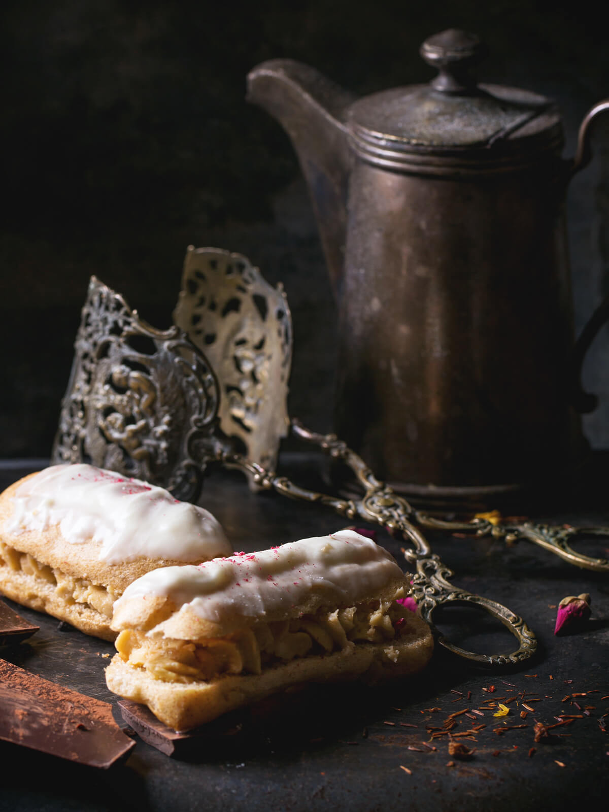 Zwei Éclairs mit weißer Schokoladenglasur auf dunklem Teller, im Hintergrund eine antike Kaffeekanne
