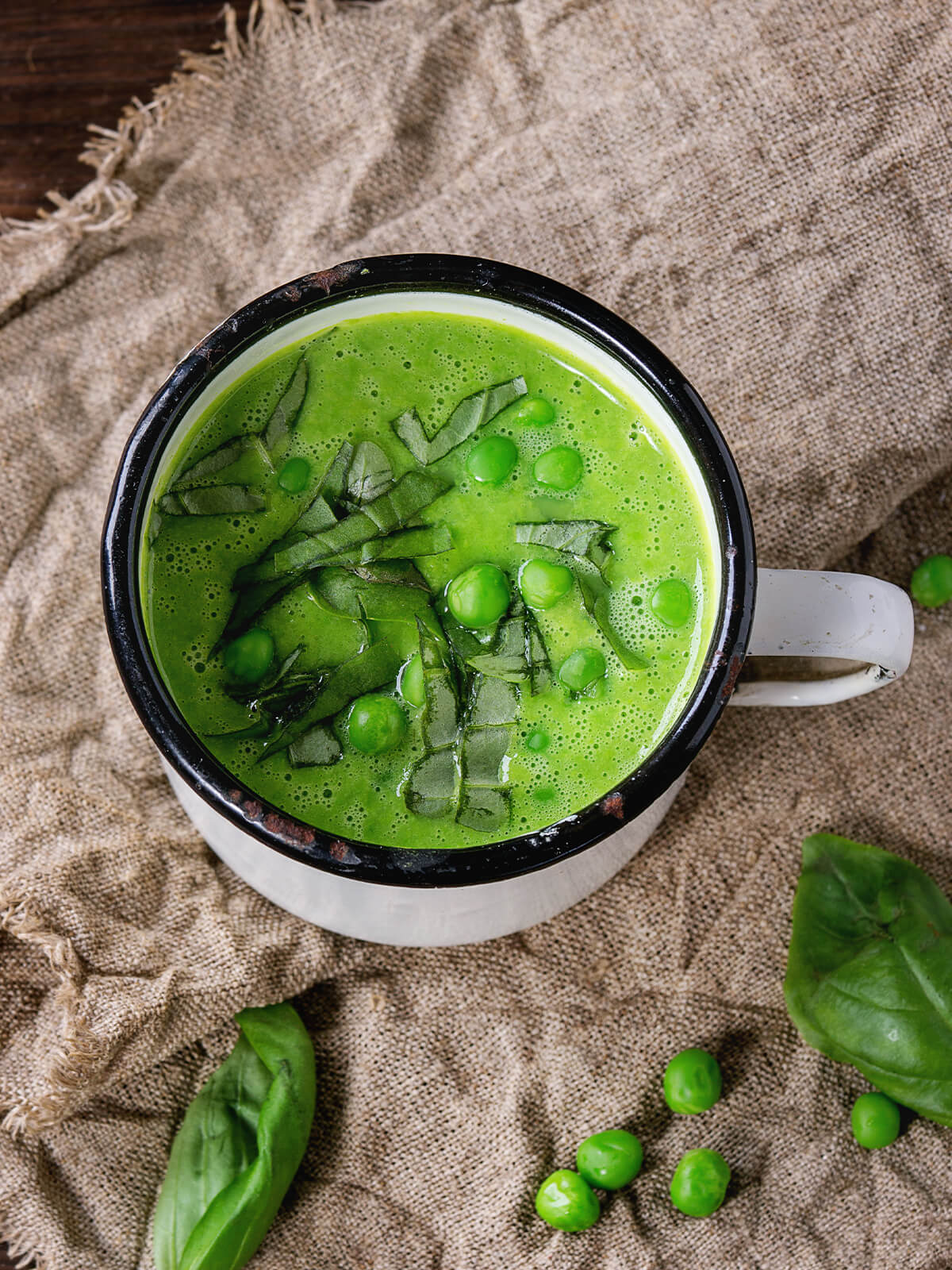 Cremige grüne Erbsensuppe in Emaille-Becher mit Basilikum und ganzen Erbsen