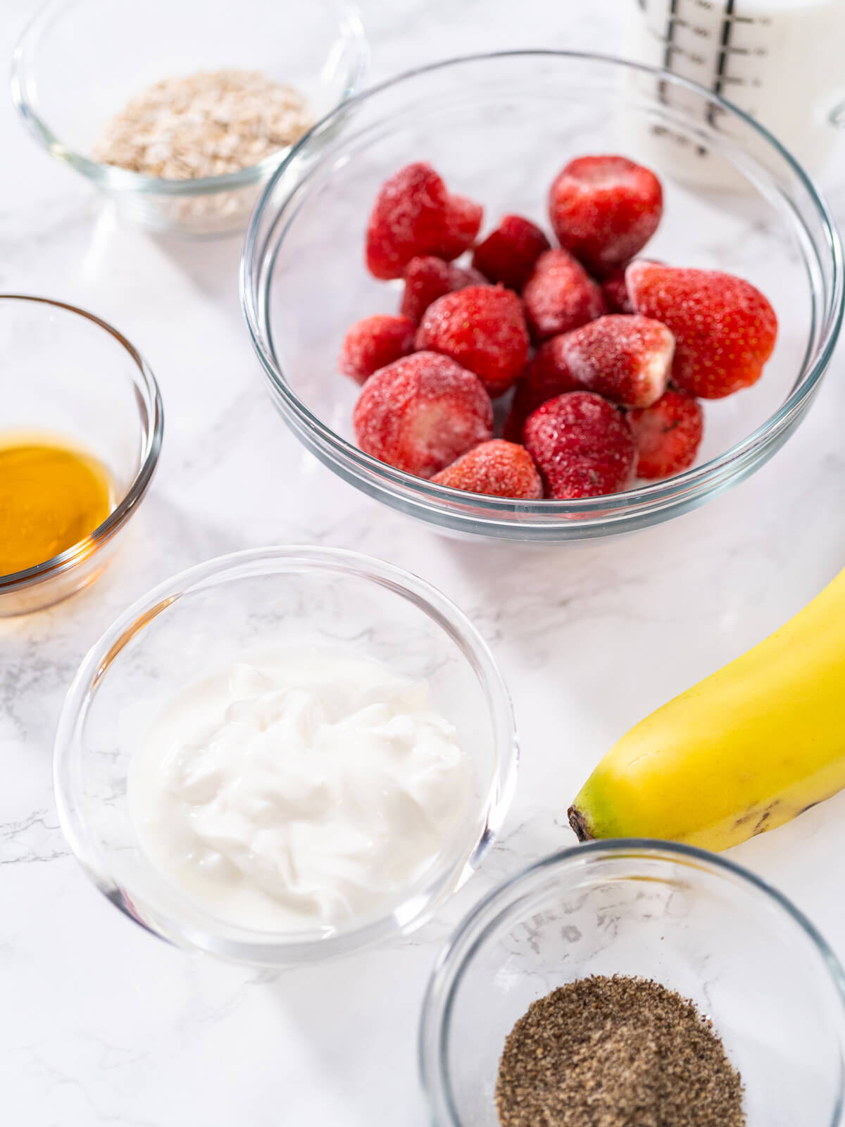 Zutaten für den Erdbeer-Bananen-Power-Smoothie in Glasschüsseln – feine Haferflocken, gemahlene Chiasamen, Joghurt, Ahornsirup, gefrorene Erdbeeren und eine reife Banane