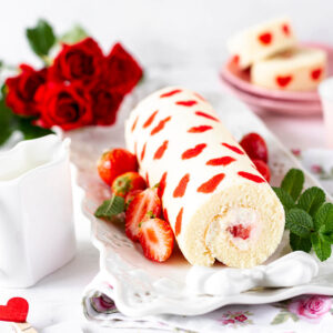 Erdbeer-Biskuitrolle mit roten Herzen im Biskuit auf weißer Servierplatte mit frischen Erdbeeren, Minze und roten Rosen