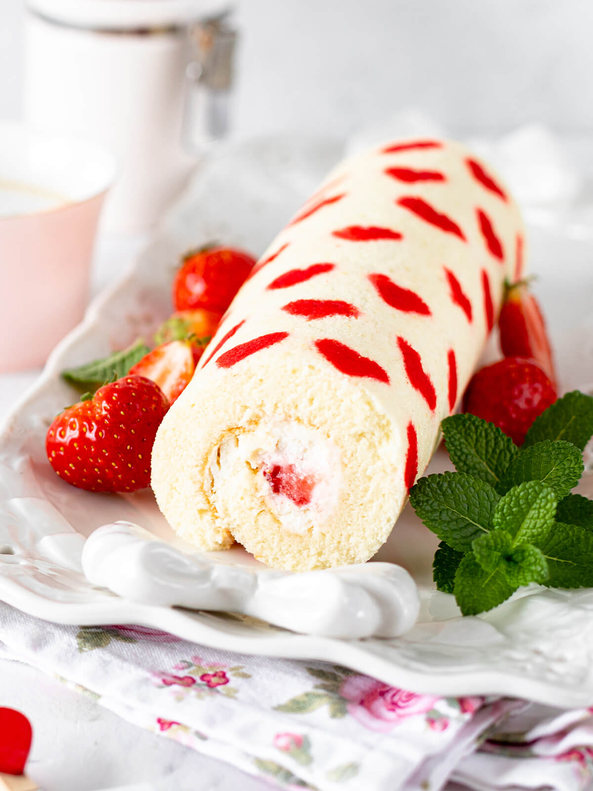 Ganze Erdbeer-Biskuitrolle mit roten Herzen im Biskuit, frischen Erdbeeren und Minze auf weißer Platte
