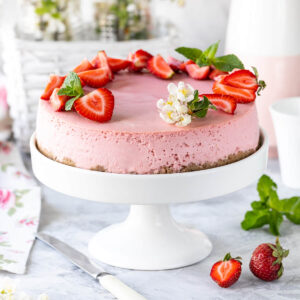 Rosa No-Bake-Erdbeer-Cheesecake ganz auf weißer Tortenplatte mit halbierten Erdbeeren