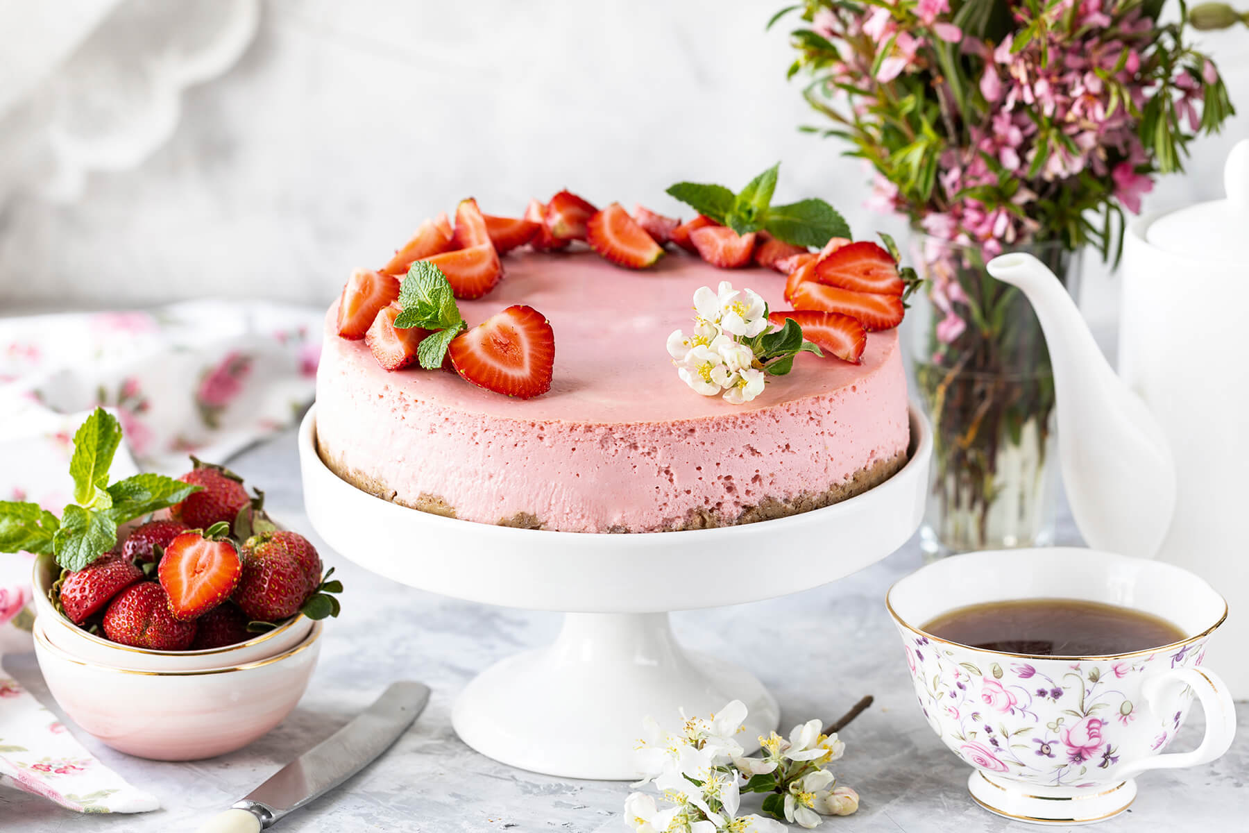 Rosa No-Bake-Erdbeer-Cheesecake mit halbierten Erdbeeren und Blüten auf weißer Tortenplatte