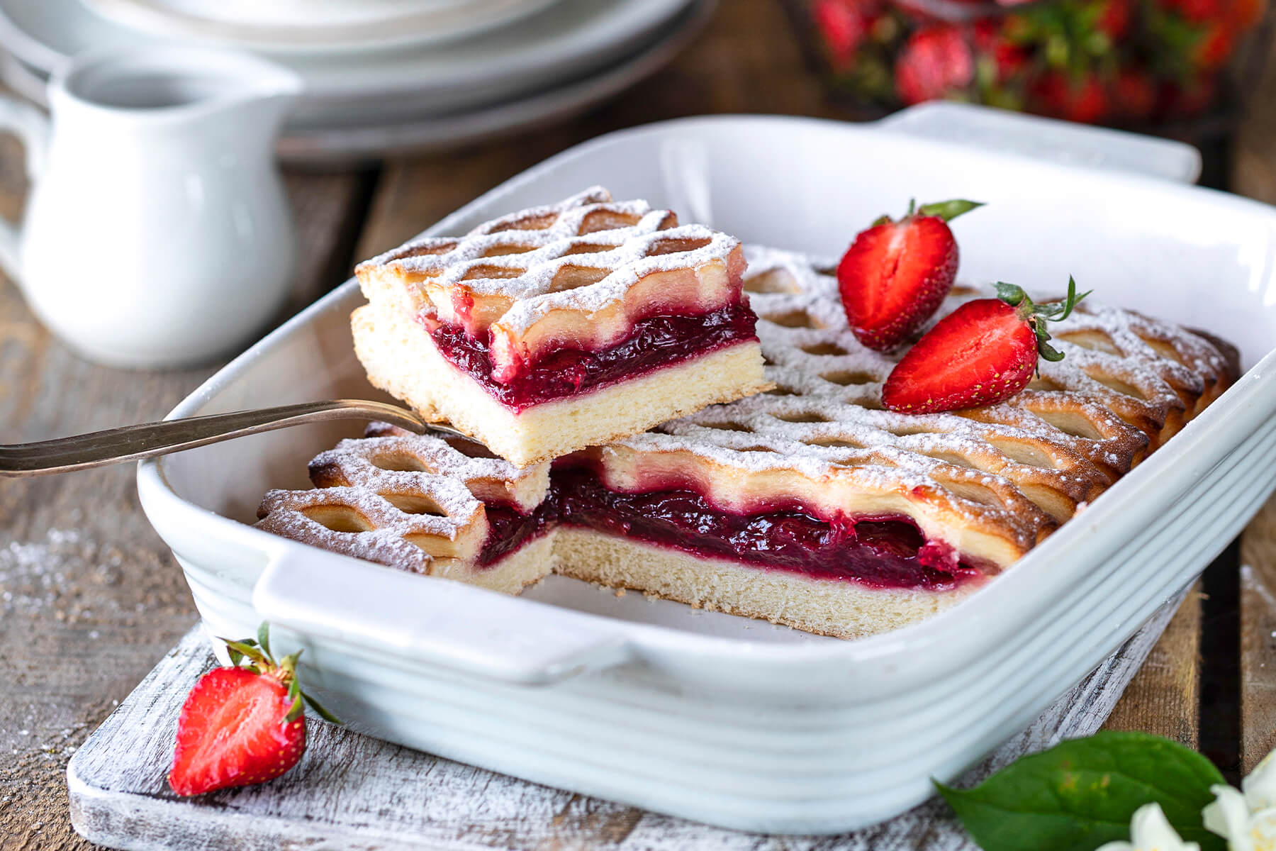 Zwei Stücke Erdbeer-Gitterkuchen in der Form, ein Stück angehoben, mit sichtbaren Schichten aus Hefeteig und Erdbeerfüllung