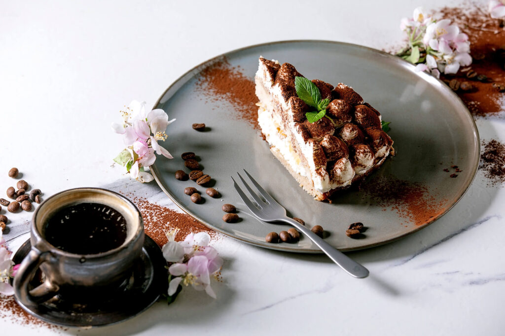 Stück glutenfreie Tiramisu-Torte mit Minzblatt auf grauem Teller, daneben eine Tasse Espresso
