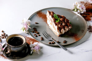 Stück glutenfreie Tiramisu-Torte mit Minzblatt auf grauem Teller, daneben eine Tasse Espresso