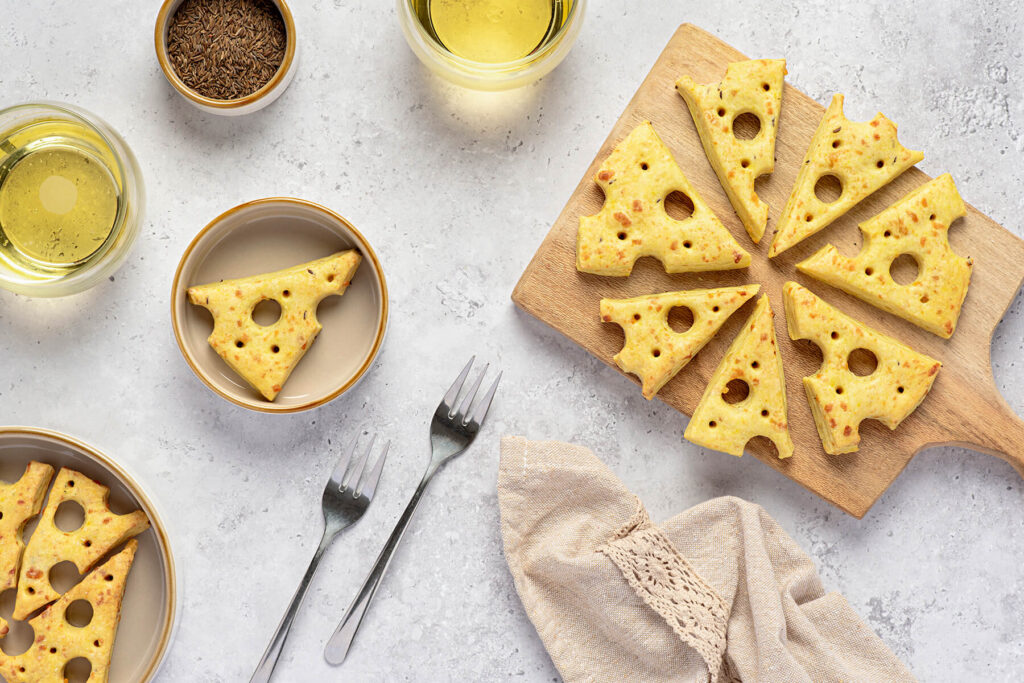Käse-Cracker in Emmentaler-Form auf einem Holzbrett im Kreis angeordnet, mit orangeroten Sprenkeln und einzelnen Kümmelkörnern auf der glänzenden Oberfläche