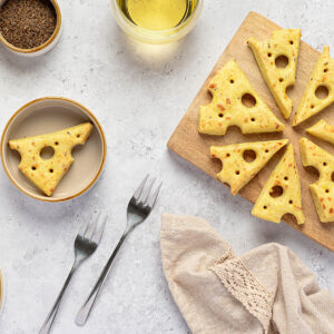 Käse-Cracker in Emmentaler-Form auf einem Holzbrett im Kreis angeordnet, mit orangeroten Sprenkeln und einzelnen Kümmelkörnern auf der glänzenden Oberfläche