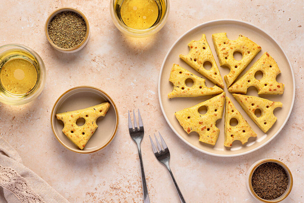 Acht Käse-Cracker in Emmentaler-Dreiecksform im Kreis auf einem hellen Teller, daneben Schälchen mit Kümmel und getrockneten Kräutern